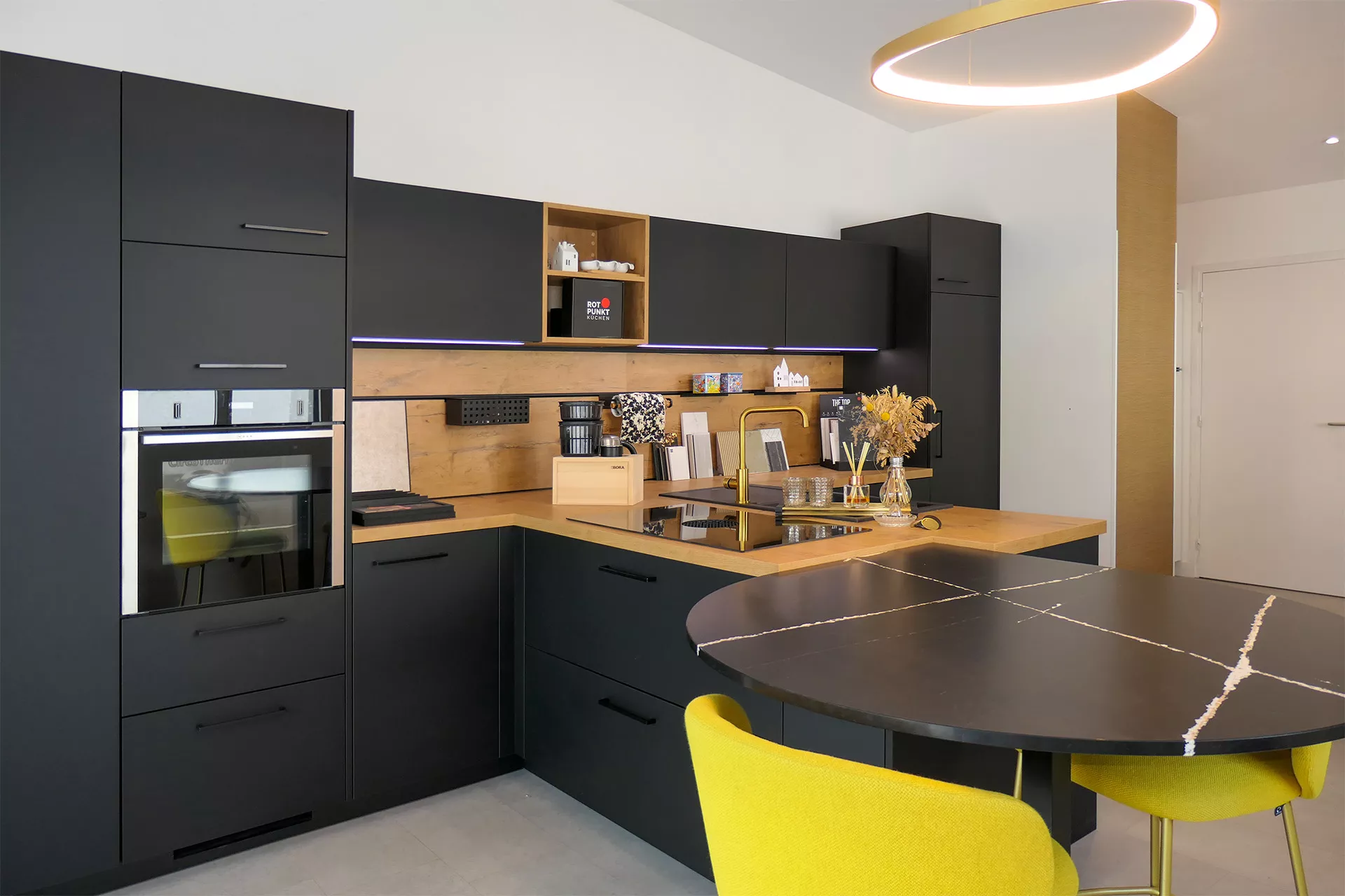 Cuisine moderne haut de gamme avec meubles noirs mats, plan de travail en bois clair, électroménager encastré, robinet doré et éclairage LED intégré. Espace repas convivial avec table ronde noire effet marbre et chaises jaunes design. Aménagement fonctionnel et élégant, idéal pour inspiration décoration intérieure et rénovation de cuisine contemporaine.