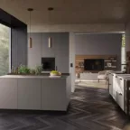 Cuisine contemporaine avec îlot central gris mat, plan de travail en bois, parquet en chevrons, meubles vitrés et grandes baies vitrées offrant une vue sur un jardin verdoyant.