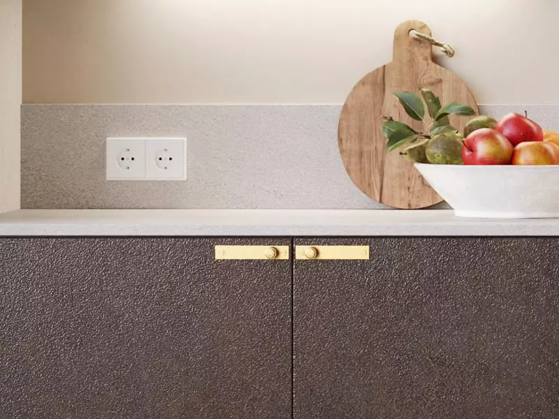 Gros plan sur un plan de travail clair avec crédence assortie, poignées dorées sur des façades texturées foncées, planche à découper en bois et coupe de fruits colorés.