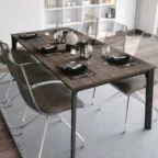 Table à manger contemporaine en bois foncé et pieds métalliques, dressée pour six convives avec vaisselle élégante et chaises transparentes, sur un tapis gris clair.