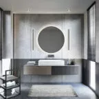 Salle de bain contemporaine avec un grand miroir rond rétroéclairé, meuble vasque suspendu gris, murs en béton ciré et lumière naturelle filtrée par des stores vénitiens.