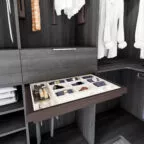 Gros plan sur un tiroir compartimenté dans un dressing en bois sombre, contenant des montres, bracelets et accessoires, sous des chemises suspendues.