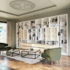 Grande bibliothèque murale blanche et bois, intégrant des rangements fermés et ouverts, des façades en cannage, dans un salon lumineux avec fauteuils verts et tables basses design sur un tapis moelleux.