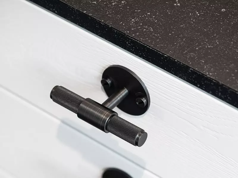 Gros plan sur une poignée de tiroir au design industriel en métal noir texturé, fixée sur un meuble blanc sous un plan de travail en granit noir.