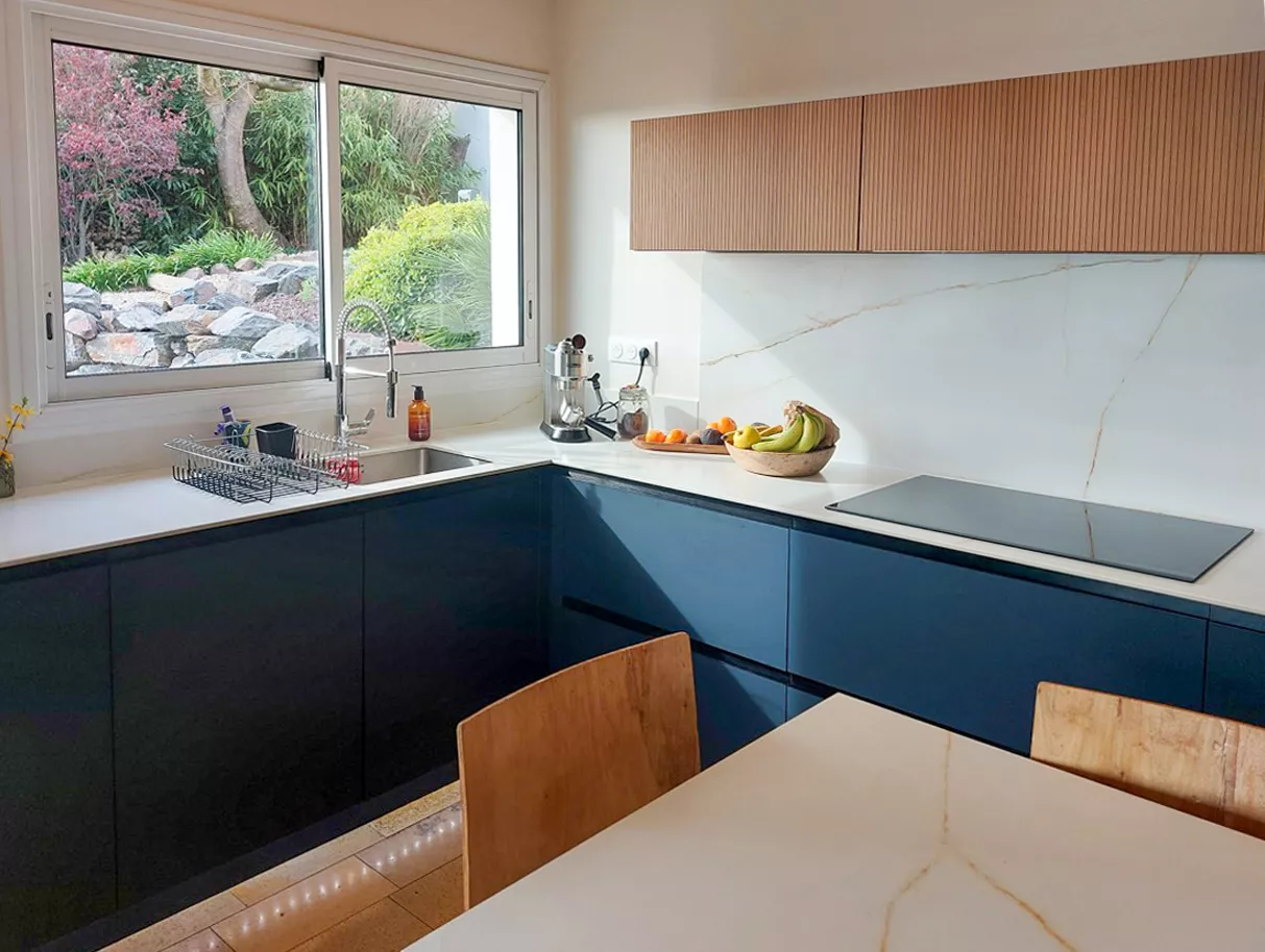 Cuisine sur mesure ouverte contemporaine réalisée sur mesure par Cuisines Premier Plan Le Havre, avec plan de travail blanc, meubles bas bleu foncé, évier sous une grande fenêtre donnant sur un jardin paysager.