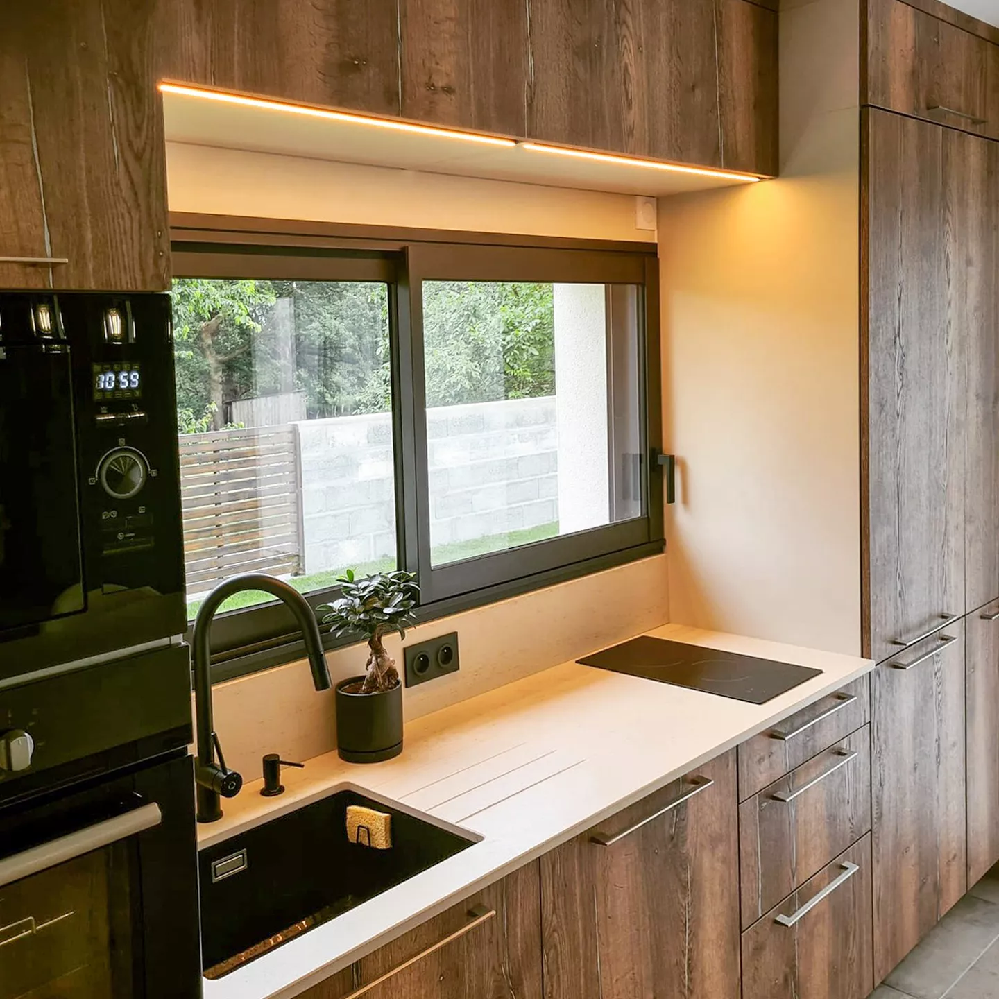 Cette vue latérale dévoile un subtil jeu de lumière grâce à la bande LED sous les meubles hauts, soulignant le plan de travail en quartz et l’électroménager encastré. L’association du bois et des finitions noires crée un design raffiné et intemporel.