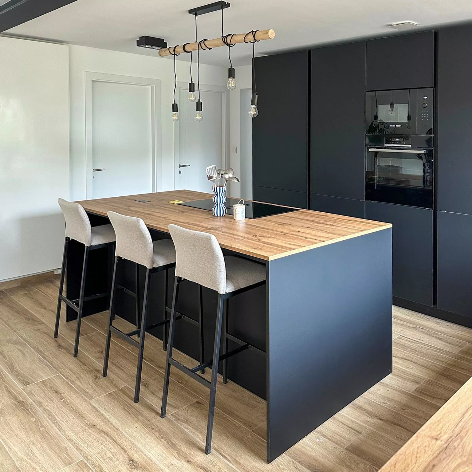 Cuisine design noire avec ilot central, chaises hautes, plancher bois, rangements intégrés et éclairage contemporain, projet réalisé à Longlaville par Cuisines Premier Plan.
