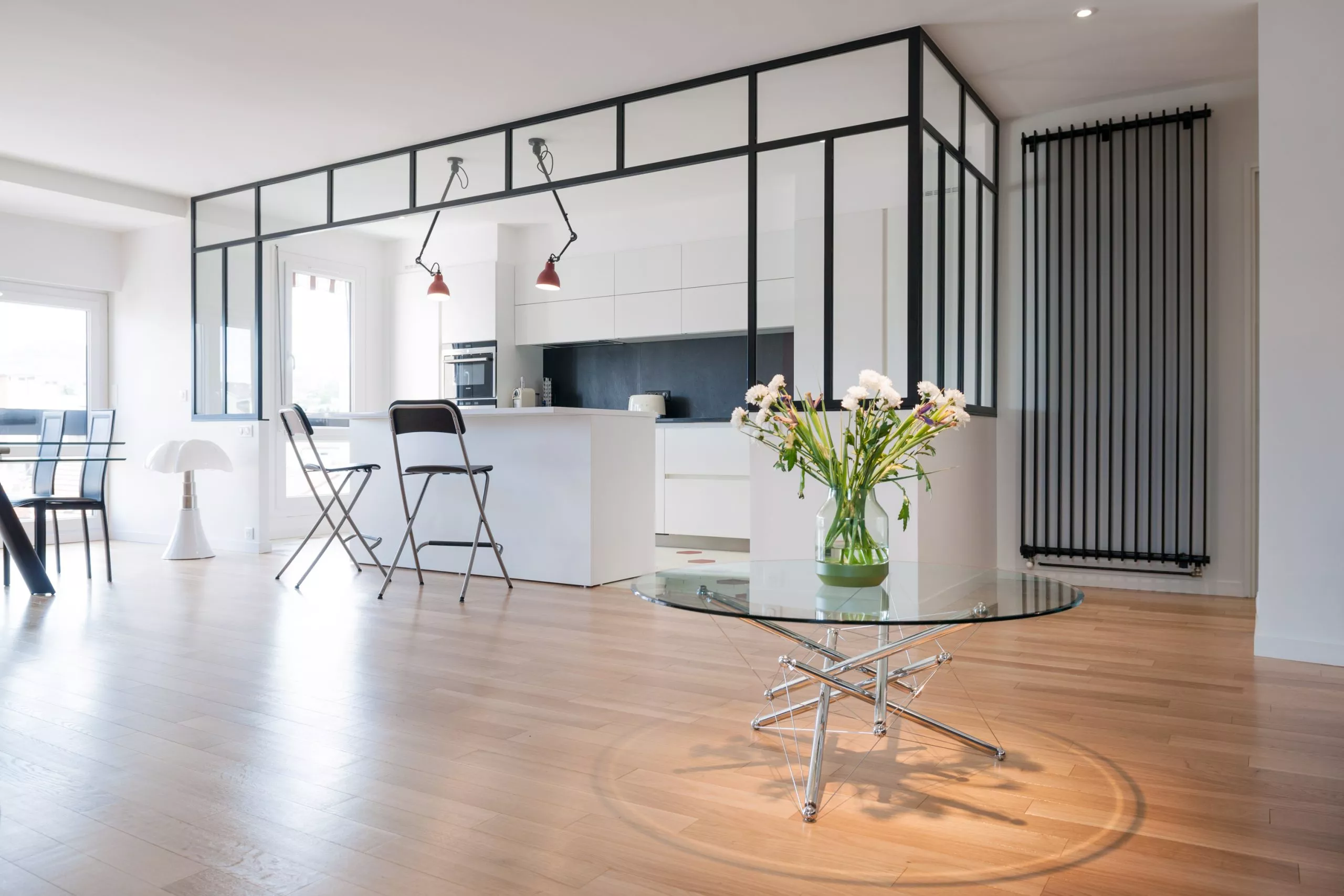 Vue d’ensemble sur une pièce à vivre baignée de lumière, où la cuisine sur mesure semi-ouverte, délimitée par une verrière, dialogue avec l’espace salon. Les parquets en bois clair, la table basse design et le radiateur vertical minimaliste soulignent l’esprit loft et chaleureux de cet intérieur