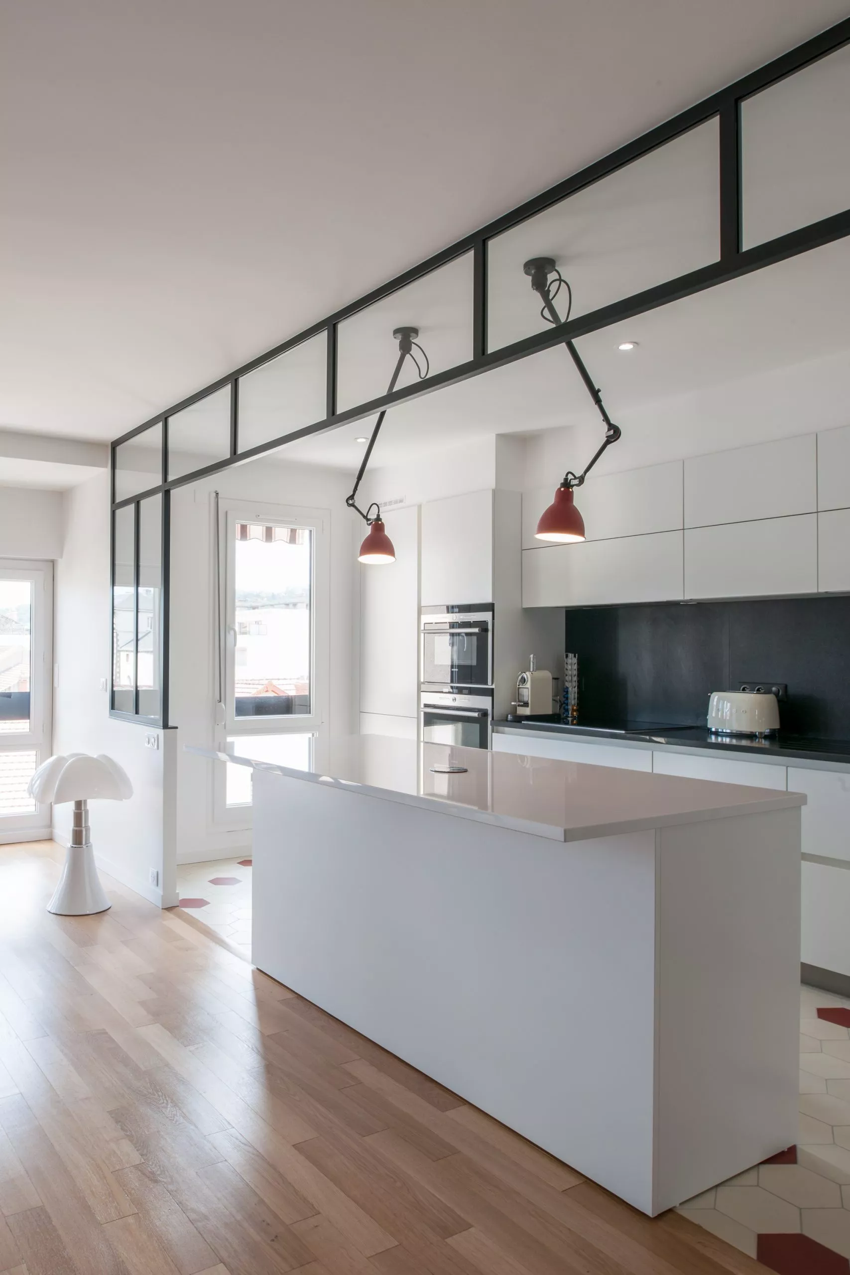 Cette cuisine ouverte bénéficie d’une luminosité exceptionnelle, grâce à de grandes fenêtres et à une verrière d’atelier qui structure subtilement l’espace sans l’alourdir. Les détails graphiques des suspensions rouges et le contraste entre les sols apportent une vraie personnalité à cette réalisation contemporaine