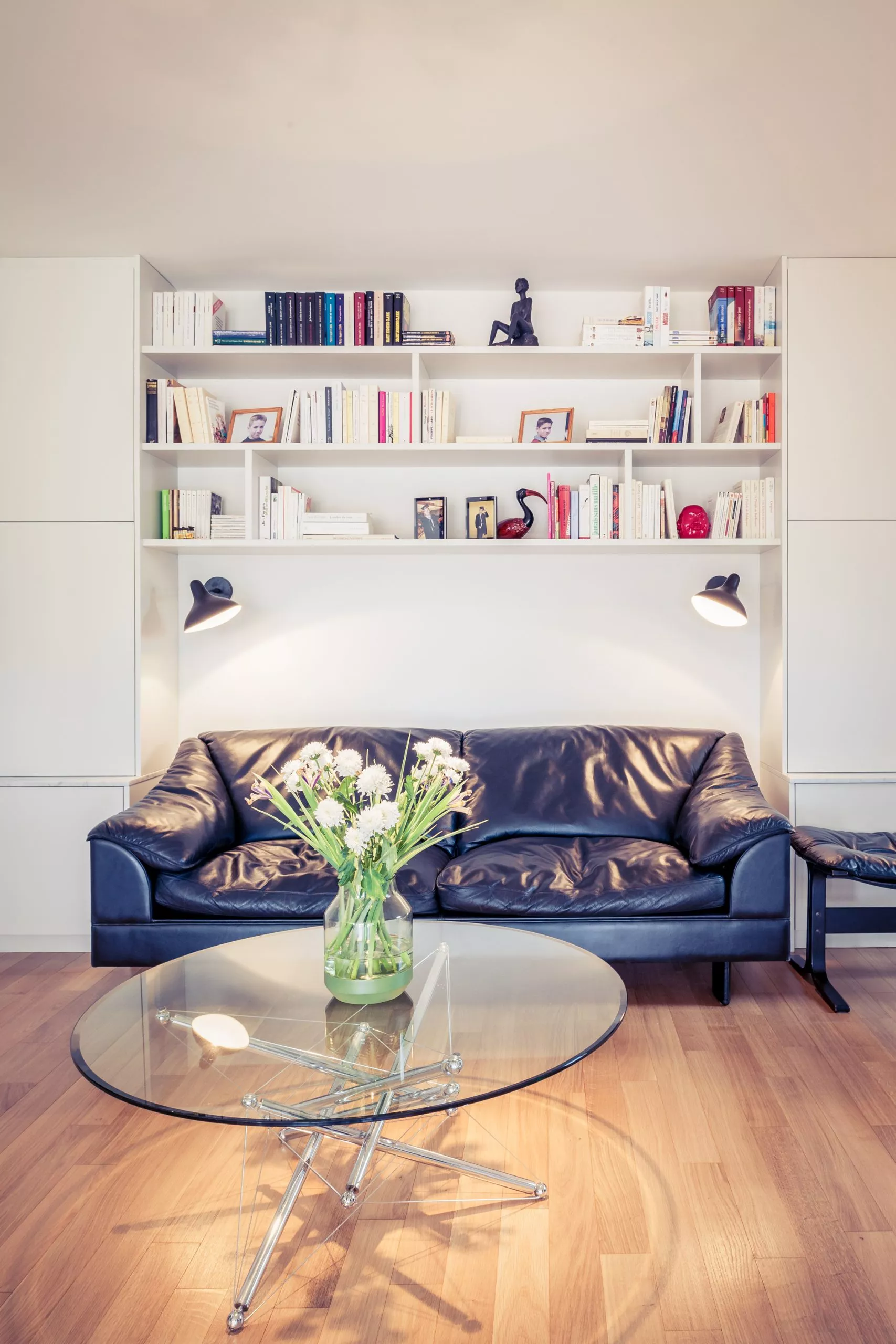 Ambiance intimiste et raffinée dans ce salon équipé d’une bibliothèque sur-mesure et d’un canapé en cuir noir. La table basse en verre et chromes renforce la luminosité et met en valeur la décoration épurée, idéale pour un espace de lecture ou de détente