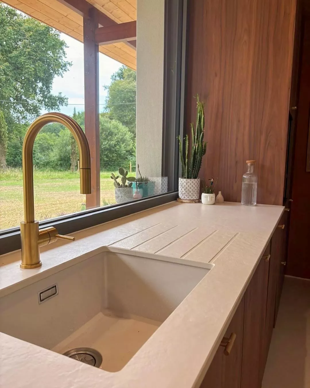 Évier encastré sous une large fenêtre avec mitigeur doré, plan de travail clair strié et meubles en bois noyer dans une cuisine moderne conçue par Premier Plan Anglet