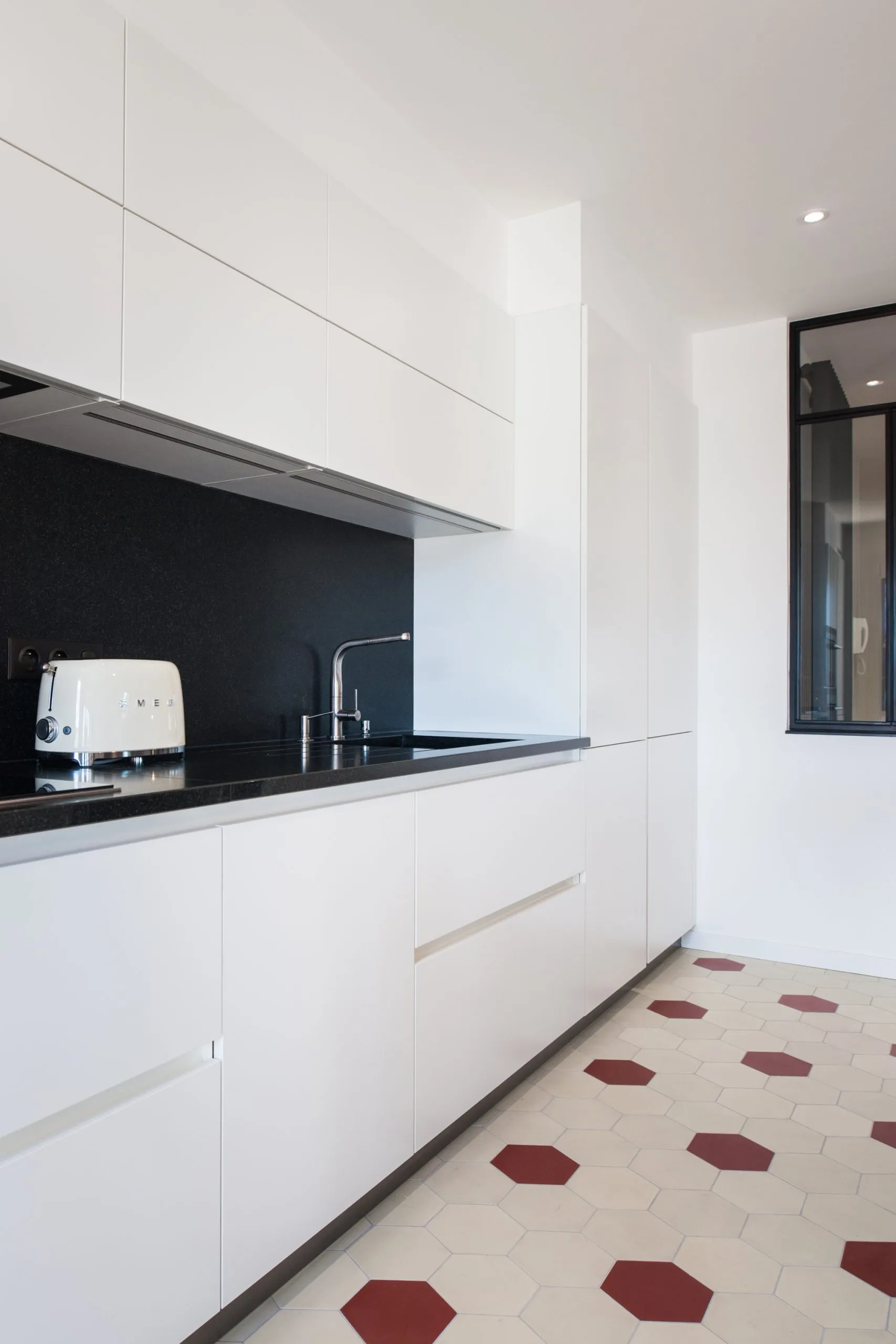 Gros plan sur le linéaire de cuisine sur mesure signé Premier Plan Cournon, avec façades blanches sans poignée, plan de travail et crédence en granit noir. L’ensemble est sublimé par un sol en carreaux de ciment hexagonaux rouges et beige et une verrière industrielle en fond de pièce.