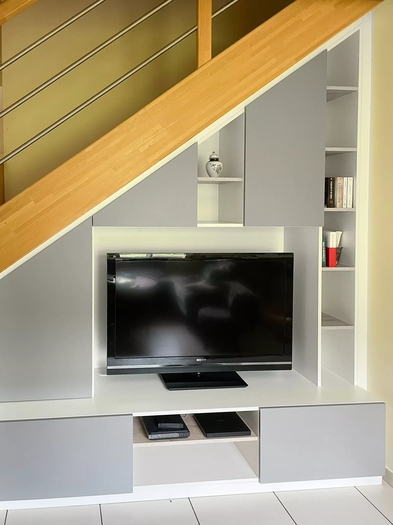Aménagement sous escalier sur mesure combinant un espace TV, des niches décoratives ouvertes et des placards fermés gris et blanc pour un salon moderne.