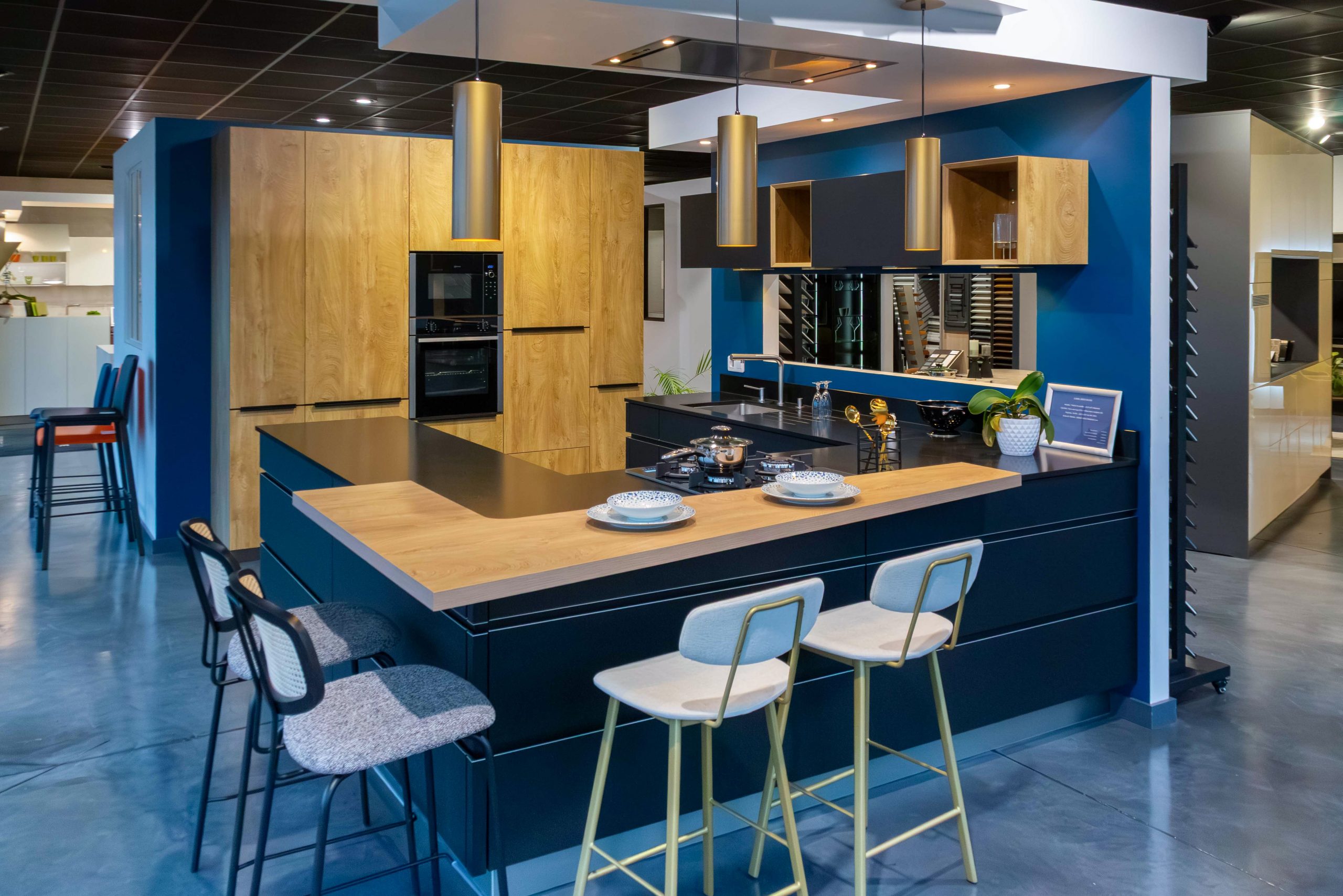 Cuisine moderne en U avec un îlot central noir mat surmonté d'un plan de travail en bois clair. Les murs sont d'un bleu canard profond, contrastant avec des colonnes de rangement en bois naturel intégrant un four encastré. Trois suspensions cylindriques dorées surplombent le bar où sont disposés des tabourets élégants. L'espace est baigné d'une lumière douce, mettant en valeur des finitions haut de gamme dans un showroom.