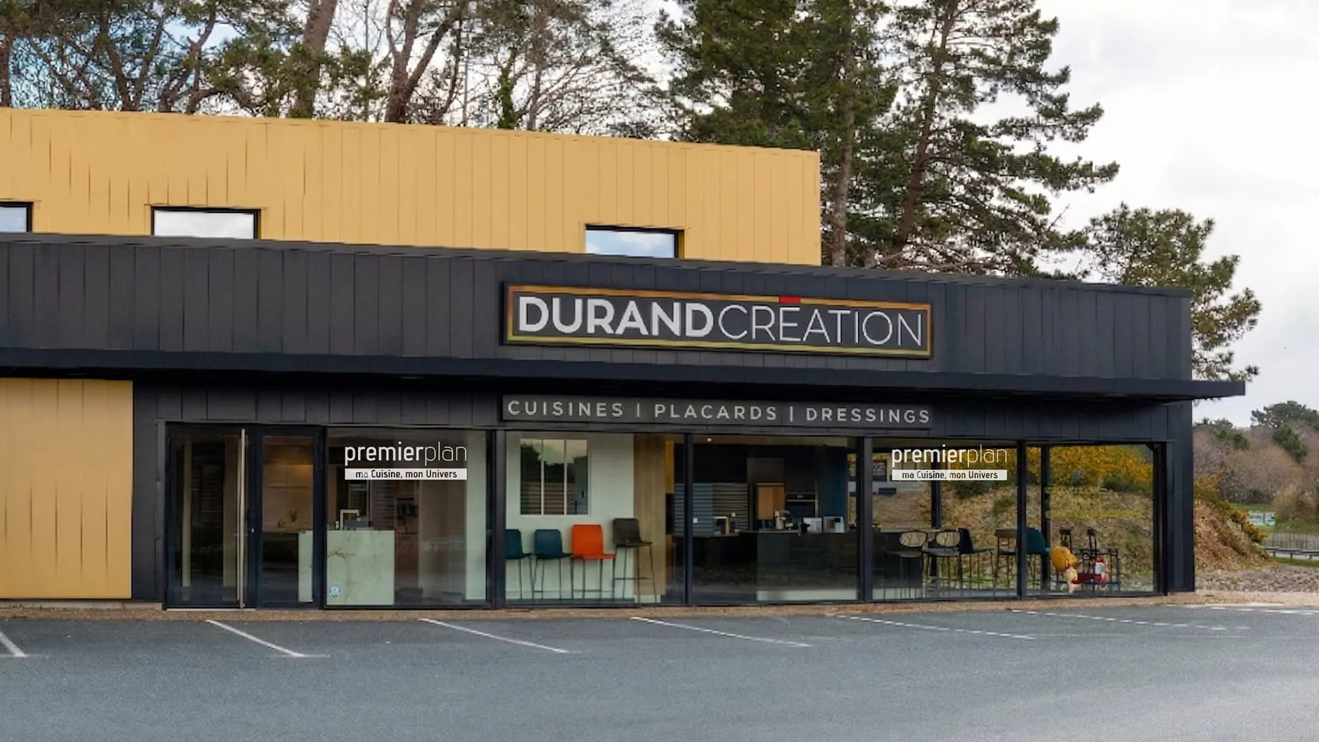 Vue extérieure d'un bâtiment commercial moderne aux façades noires et ocres. L'enseigne principale indique "DURAND CREATION" avec la mention "CUISINES | PLACARDS | DRESSINGS". De larges vitrines laissent apparaître l'intérieur d'une exposition de cuisines et de mobilier. Un fourgon blanc est stationné sur le parking bitumé devant l'entrée, avec des arbres en arrière-plan sous un ciel clair.