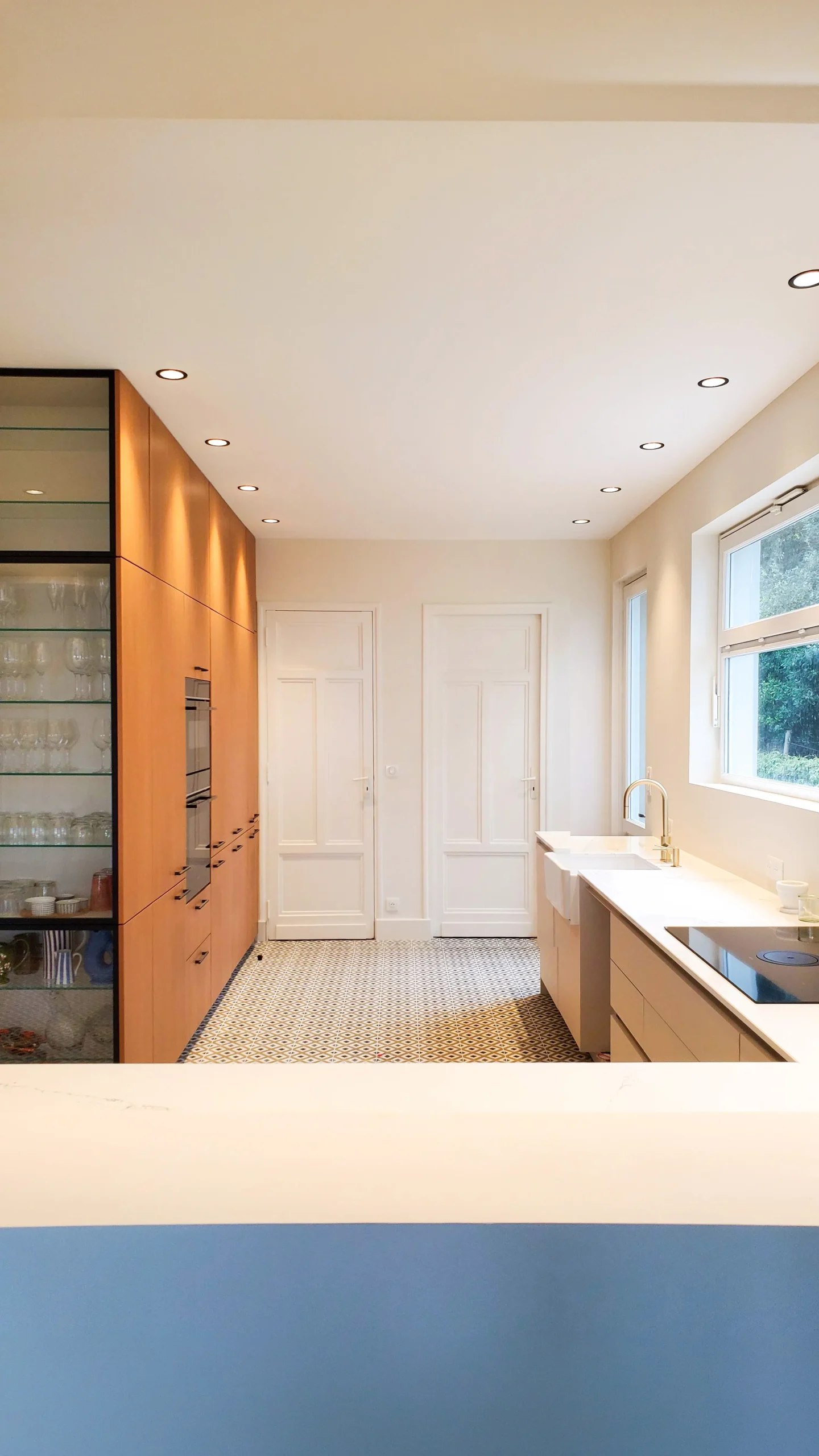 Cuisine sur mesure avec grande colonne en bois doré avec vitrine à verrerie, façades basses blanches épurées, plan de travail blanc, robinetterie dorée et carrelage ancien géométrique au sol, réalisée par Cuisines Premier Plan à Montauban