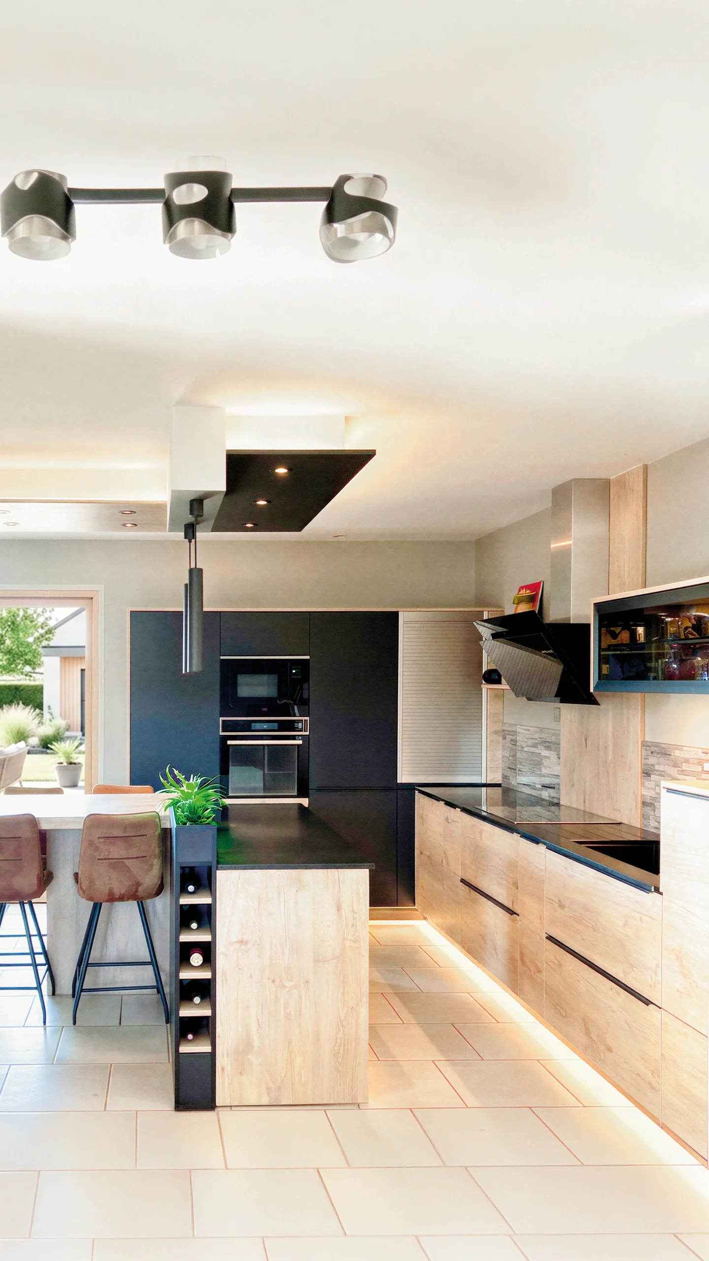Cuisine sur mesure avec façades bois clair et panneaux noirs mats, îlot central avec plan de travail noir, cave à vin intégrée dans la joue d'îlot, éclairage LED sous meubles et tabourets cuir brun, réalisée par Cuisines Premier Plan à La Ferté-Macé