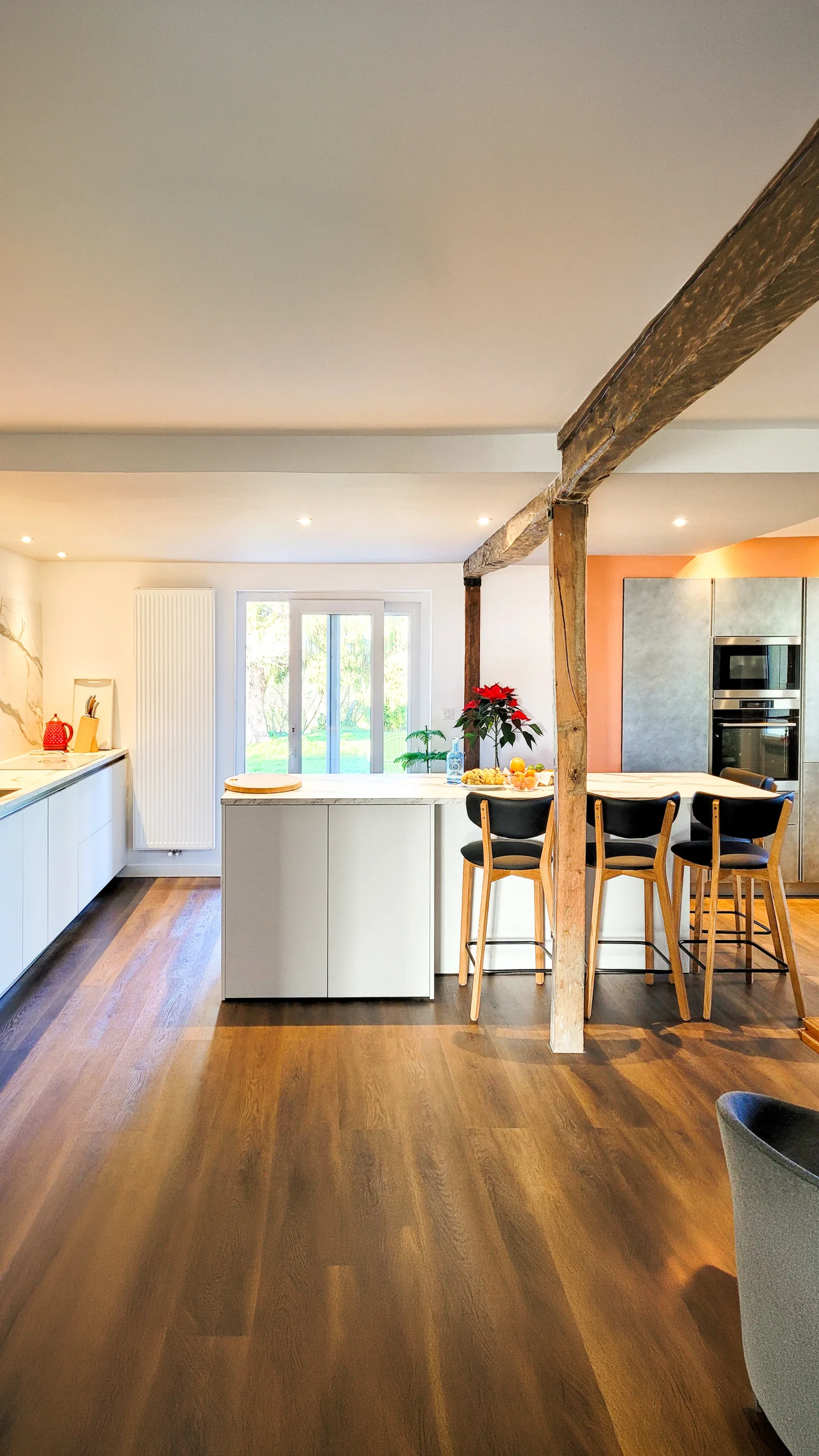 Cuisine sur mesure épurée avec façades blanches lisses, crédence marbre, colonne effet béton pour électroménager encastré, poutres apparentes en bois brut et parquet chêne foncé, réalisée par Cuisines Premier Plan à Pau