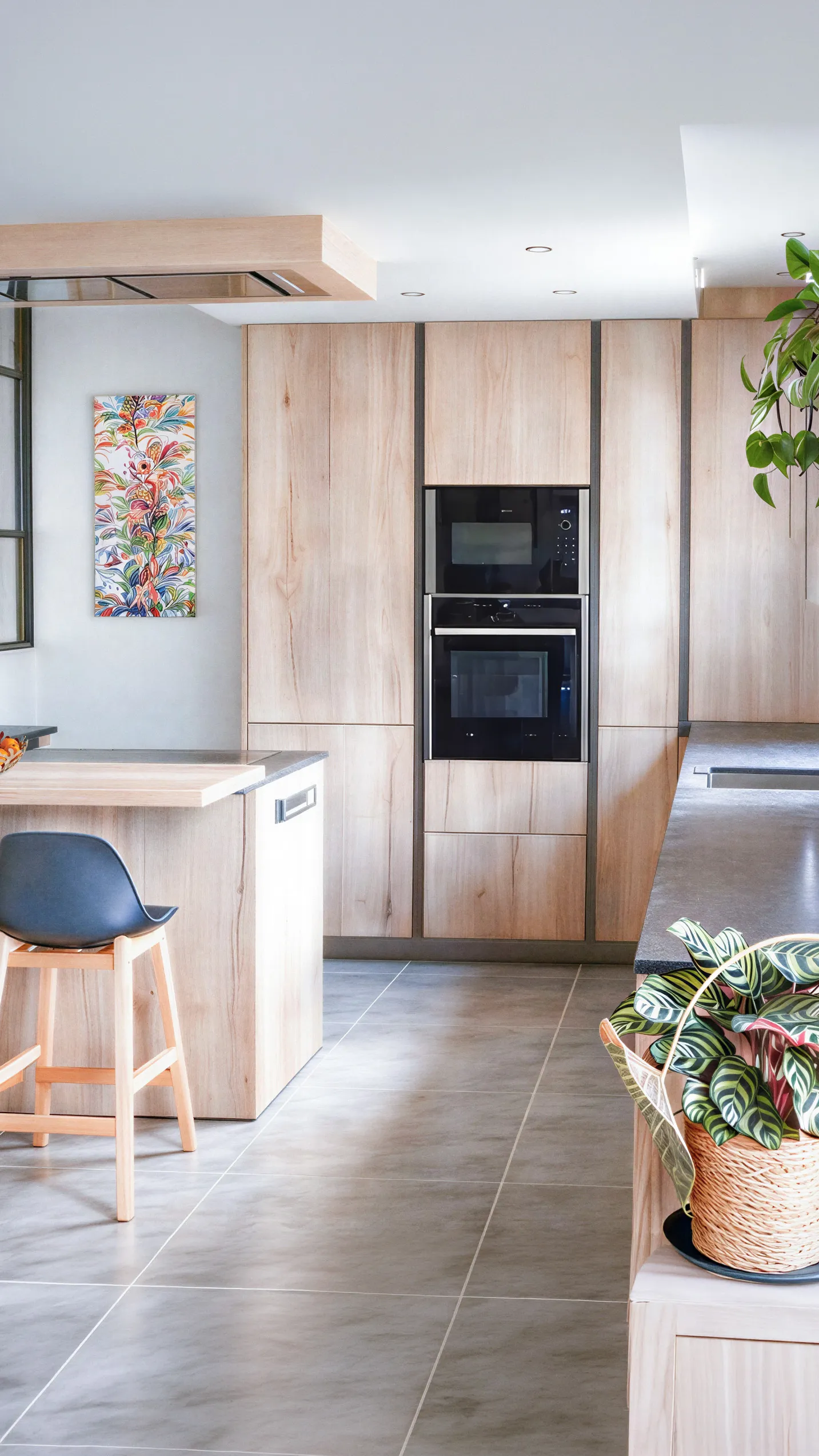 Cuisine sur mesure avec façades en bois clair texturé et listel gris anthracite, colonne four et micro-ondes encastrés, plan de travail gris et décoration végétale colorée, réalisée par Cuisines Premier Plan à La Suze-sur-Sarthe