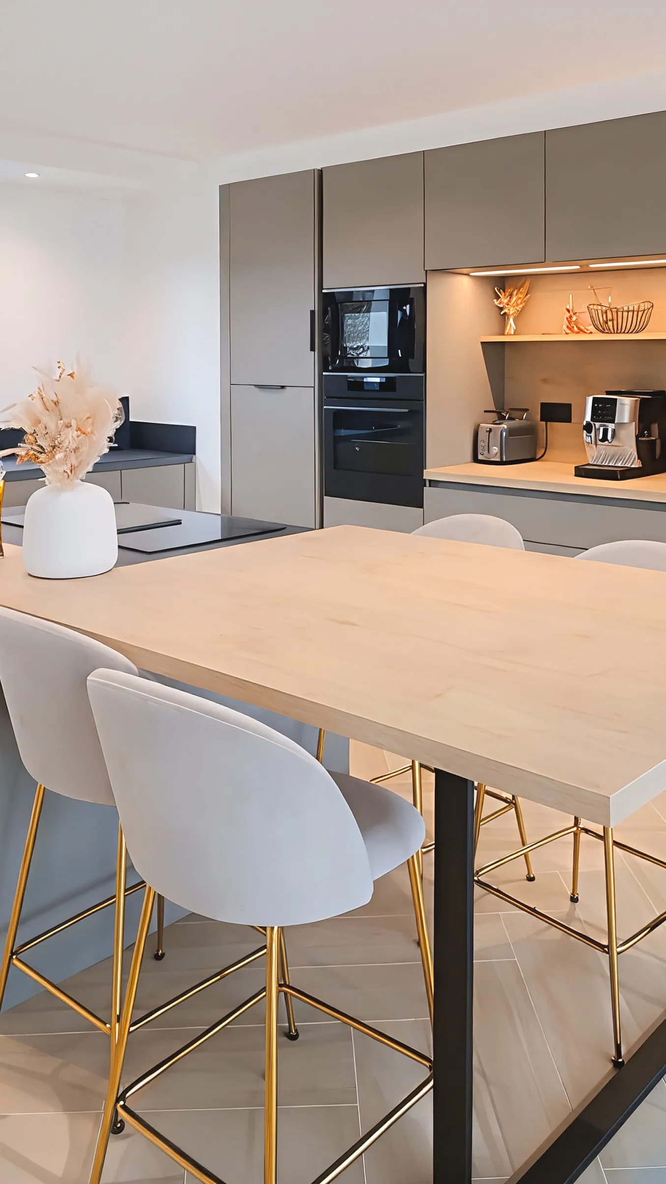 Cuisine sur mesure avec façades grises mate, angle ouvert éclairé, grand plan de travail bois clair sur pied central noir, tabourets blancs aux pieds dorés et électroménager encastré noir, réalisée par Cuisines Premier Plan à Guer