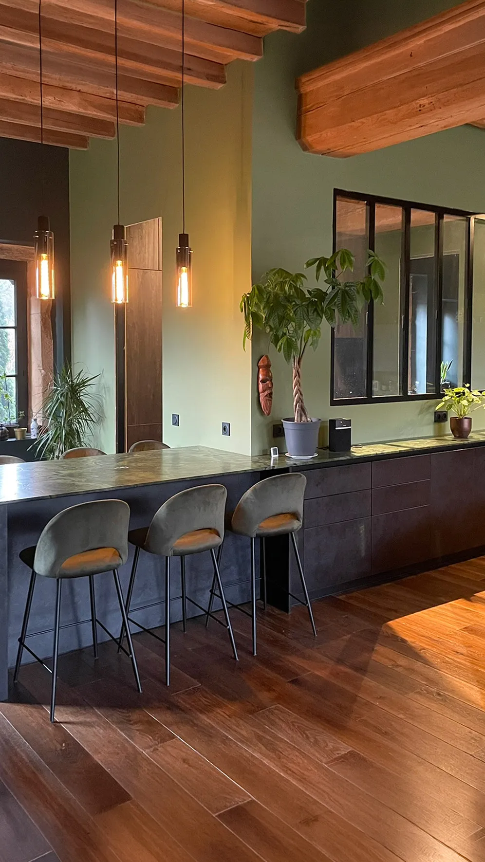 Cuisine sur mesure avec îlot bar tout noir, plan de travail en marbre vert foncé, tabourets velours gris, poutres apparentes en bois, murs vert sauge et verrière noire, réalisée par Cuisines Premier Plan au Bois-d'Oingt