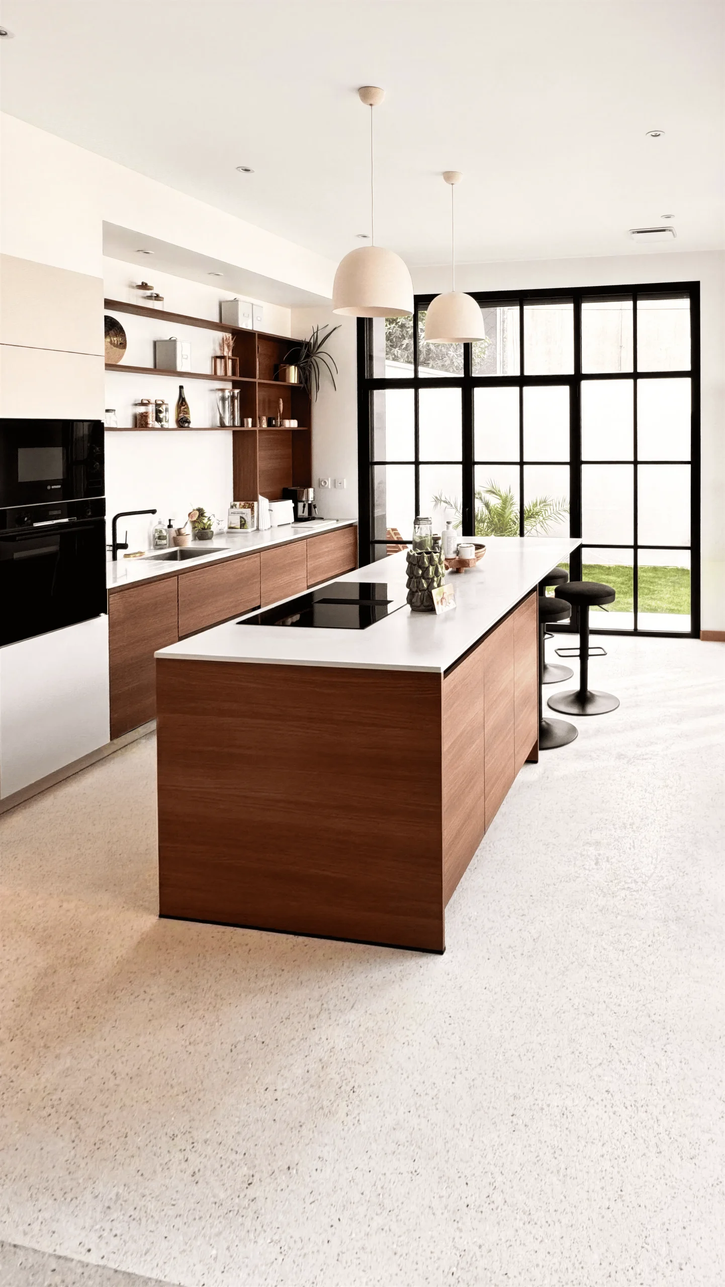 Cuisine ouverte sur mesure avec façades bois noyer chaud, plan de travail blanc, grand îlot central avec plaque de cuisson intégrée, verrière métallique noire et sol terrazzo blanc, réalisée par Cuisines Premier Plan au Havre