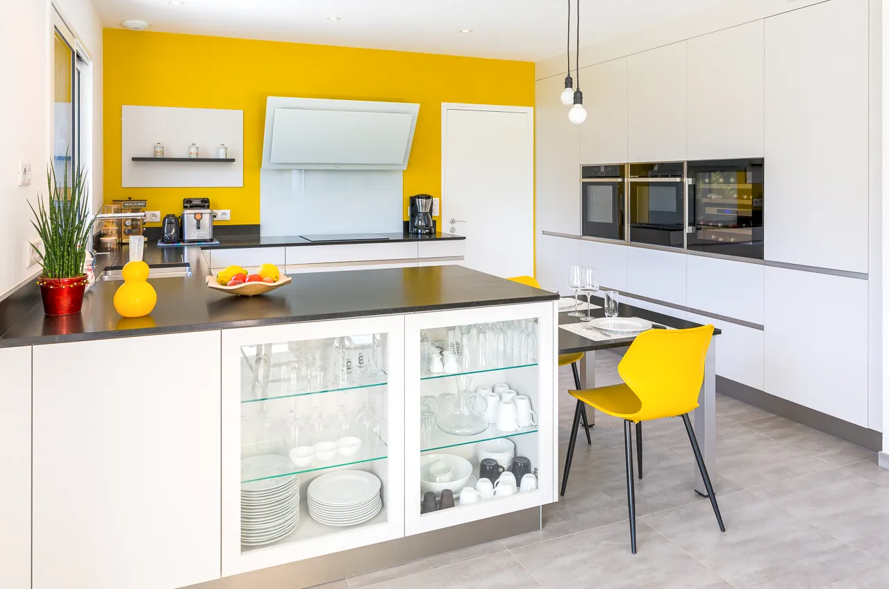 Cuisine blanche Premier Plan Pont-l’Abbé avec mur jaune, plan de travail noir, coin repas et vaisselier vitré intégré