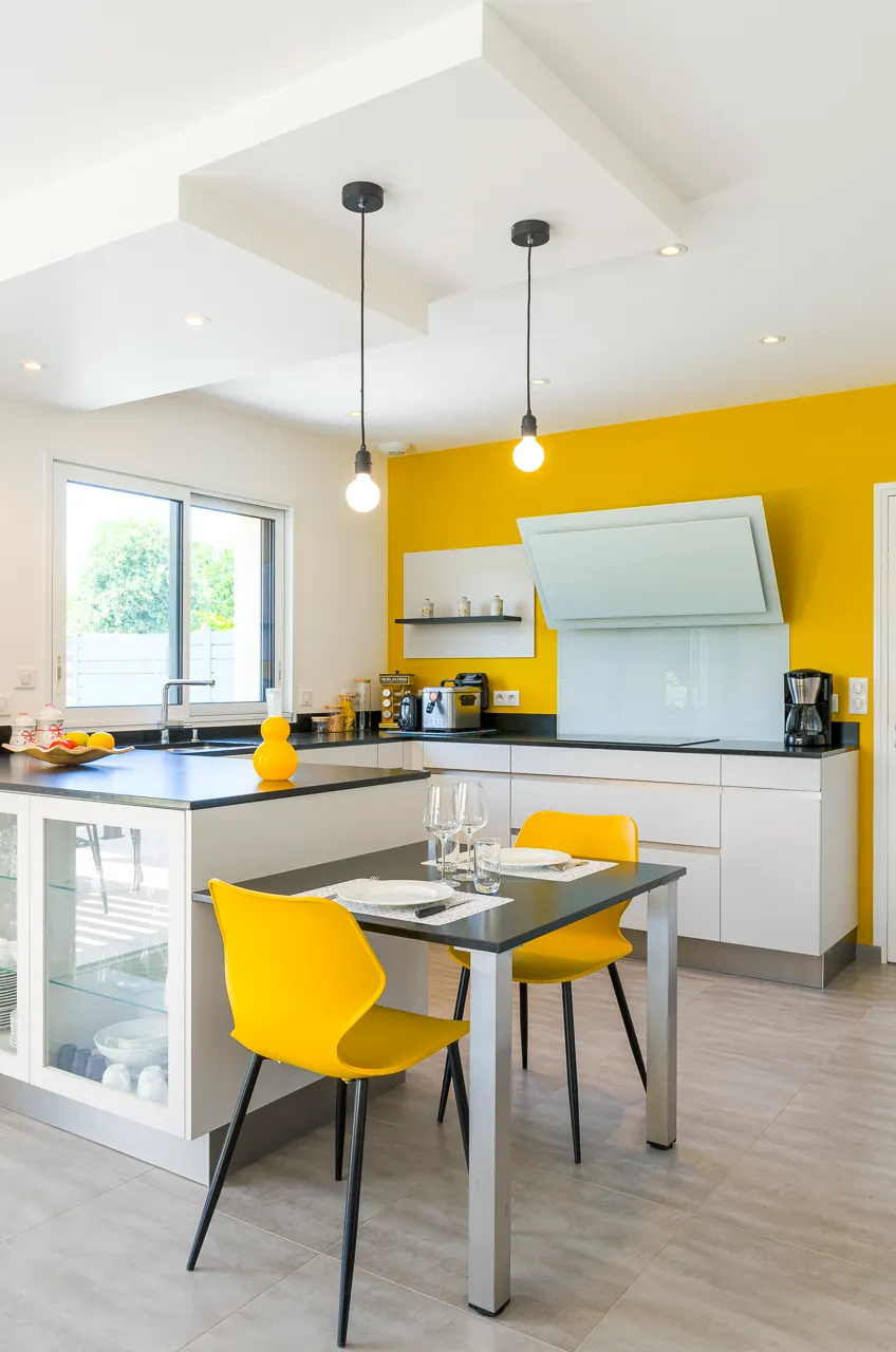 Cuisine lumineuse Premier Plan Pont-l’Abbé avec façades blanches, mur jaune, table repas noire et chaises jaunes